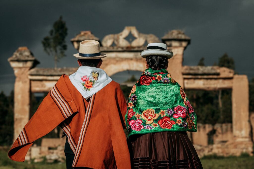 A couple in vibrant traditional clothing stands facing ancient ruins in Jauja, Perú.